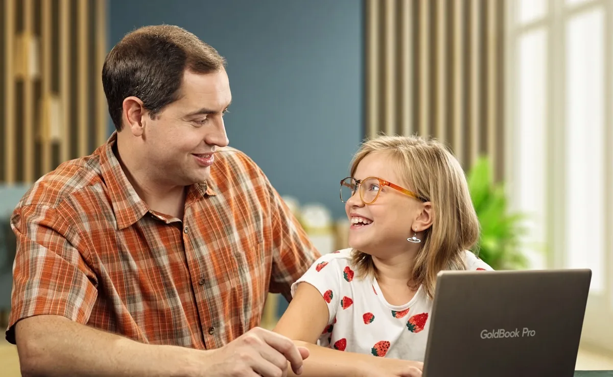 A father and his daughter smile at each other as she studies at home on a GoldBook Pro laptop